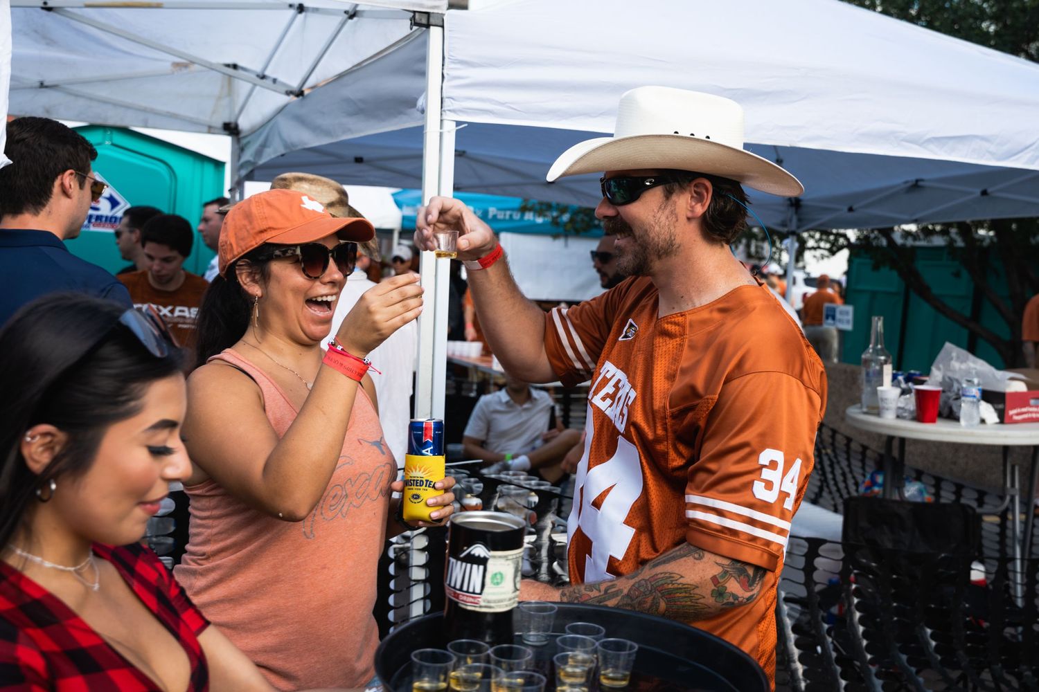 Tailgaters interact at a drink station under white tents during a sports event.