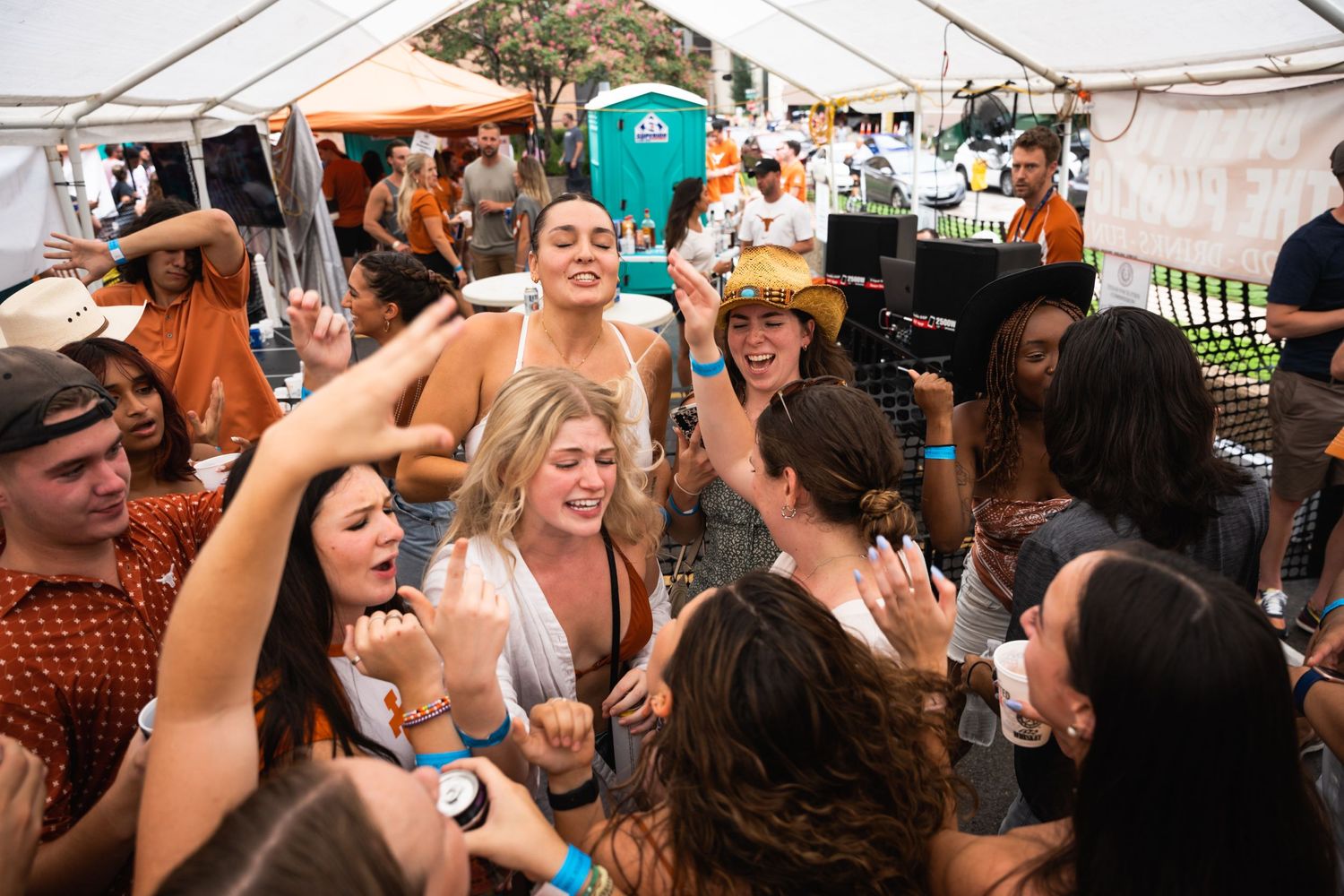 Excited crowd of partygoers celebrating under a tent with raised drinks and energetic dancing.