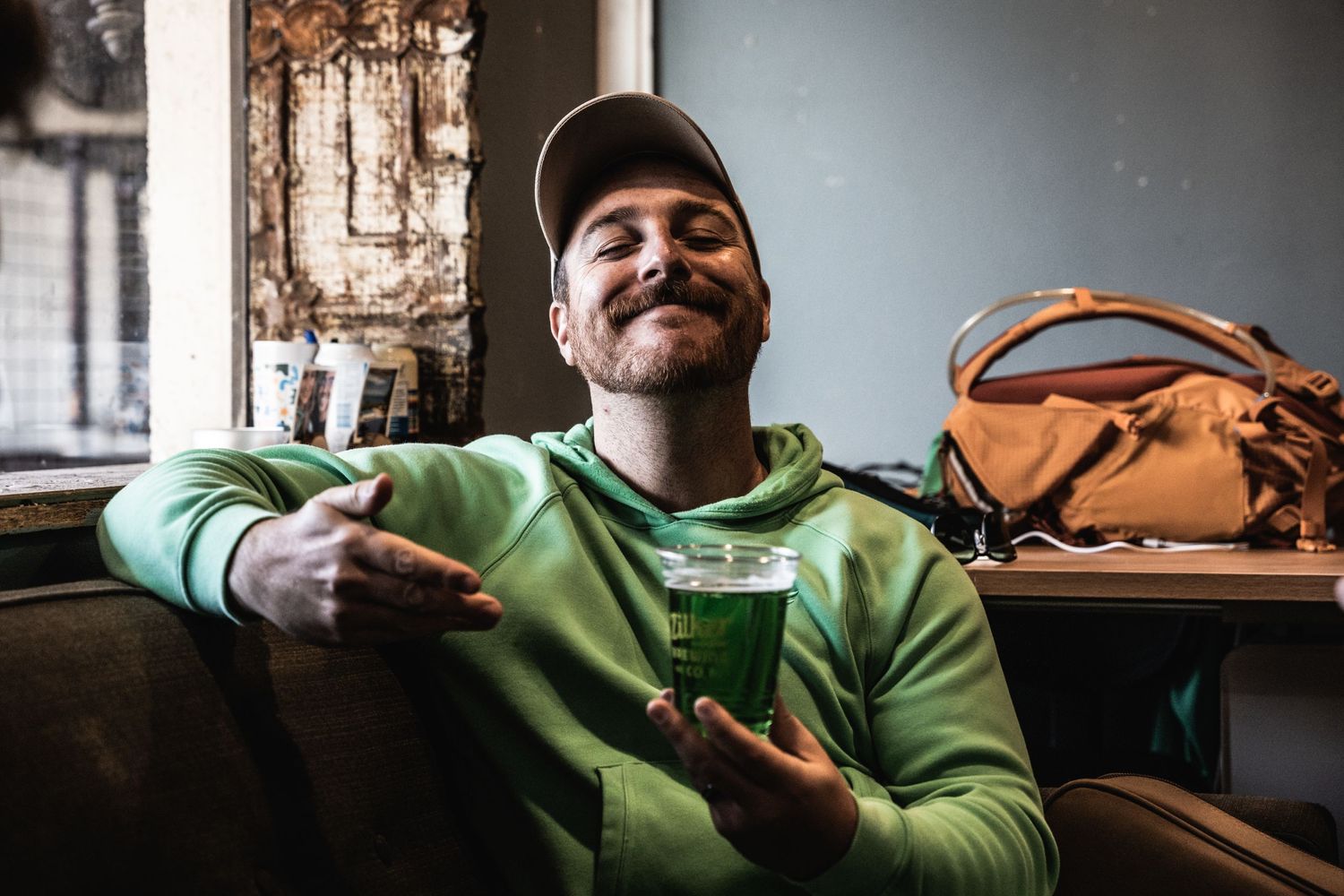 Person wearing bright green hoodie and baseball cap gestures while sitting on couch with brown bag nearby.