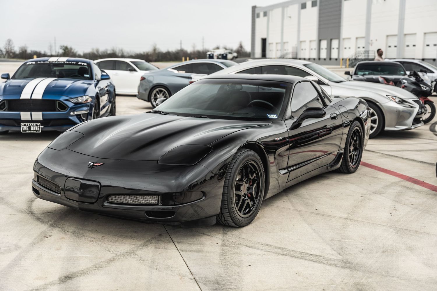 Black Chevrolet Corvette with racing stripes parked in front of industrial building.