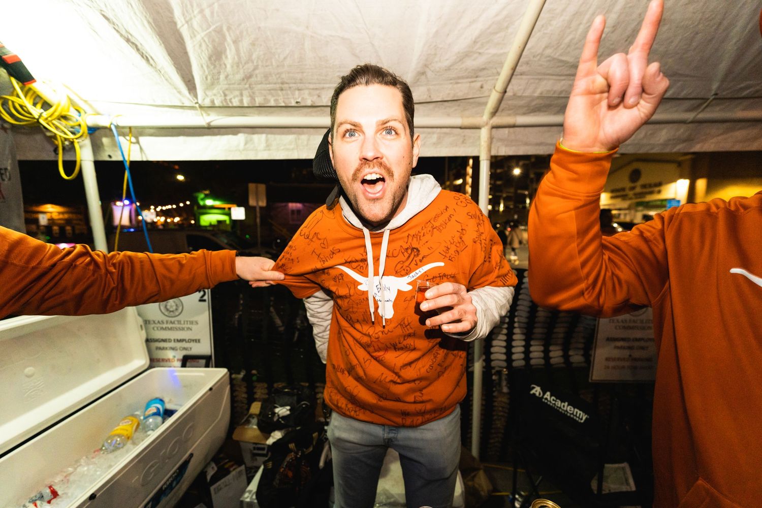 Person in orange hoodie making energetic gestures and hand signs under covered outdoor area.