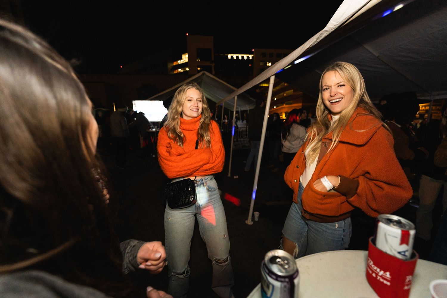 People gathered outdoors at night under tent lighting with drinks and casual conversation.
