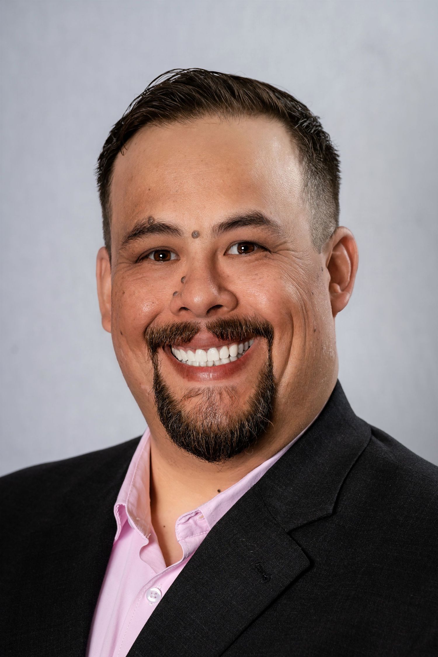 Professional headshot of person in dark suit and pink shirt with facial hair.