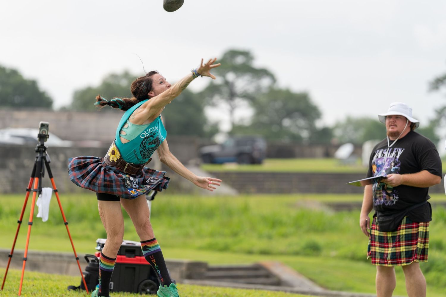 Woman in tartan skirt throws shot put at Highland Games while official in plaid kilt observes during outdoor competition.