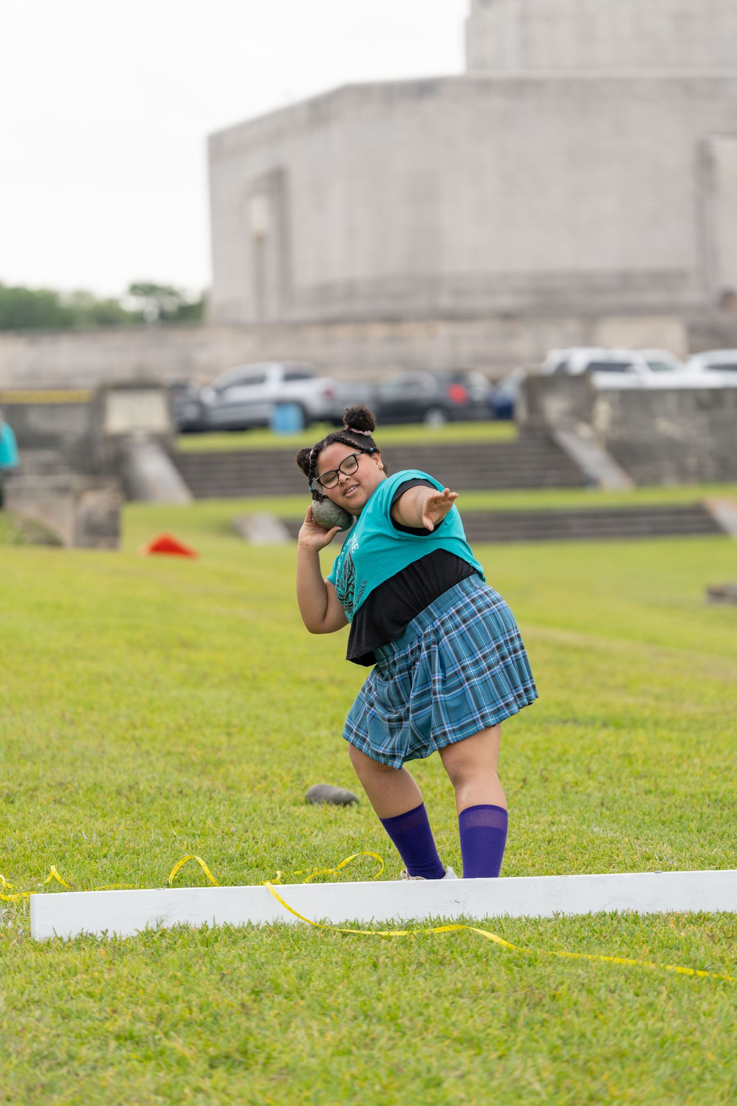 Young woman in plaid skirt and knee socks throwing shot put at outdoor Highland Games competition.