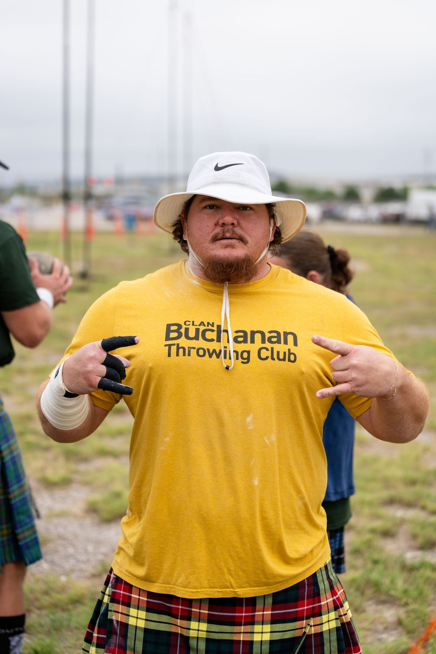 Man in yellow Buchanan Throwing Club shirt and plaid kilt posing outdoors at Highland Games competition.