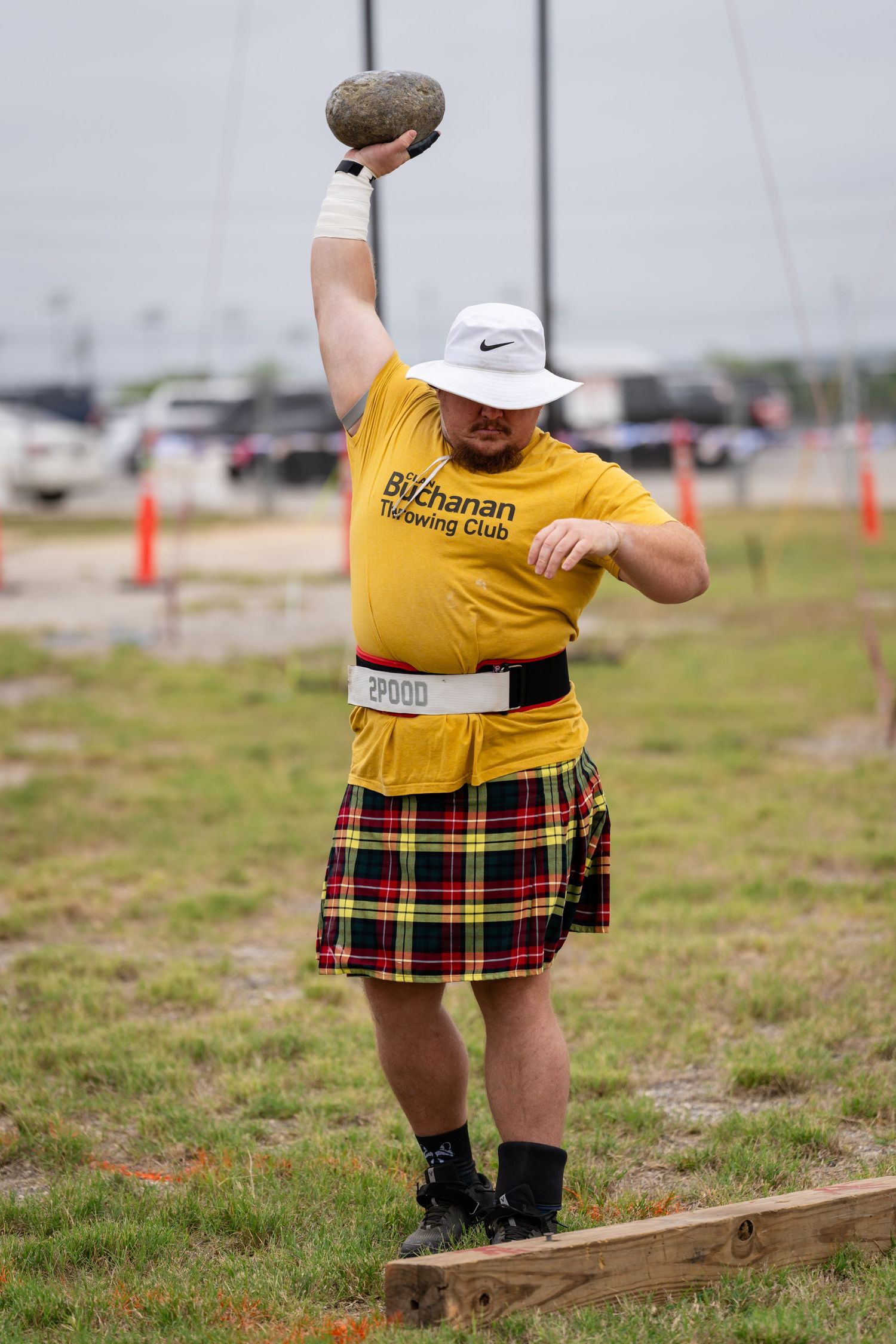 Man in yellow Buchanan Throwing Club shirt and tartan kilt raises stone overhead at Highland Games competition.