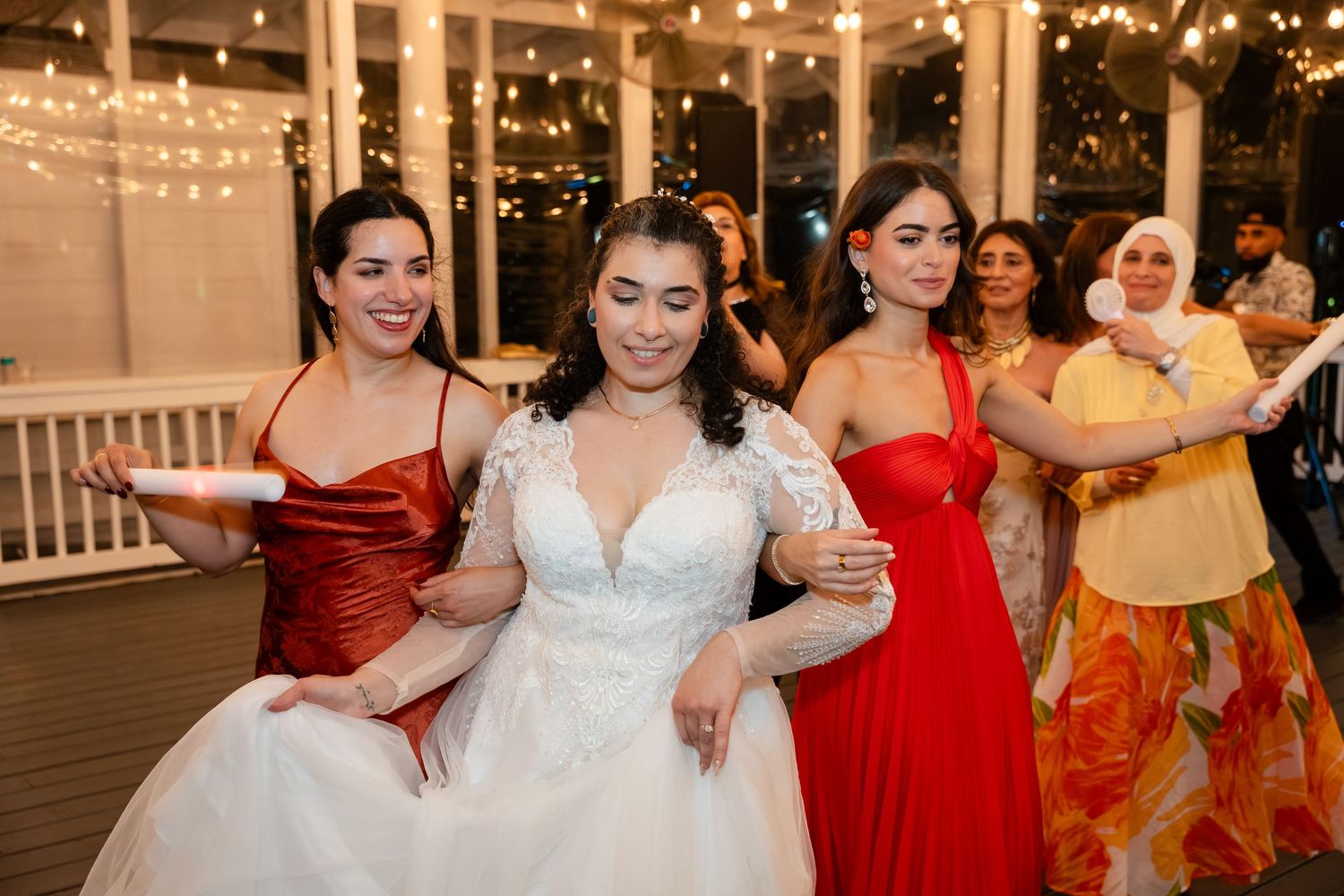 Bride in white gown dances joyfully with bridesmaids in red and colorful dresses at elegant wedding reception.