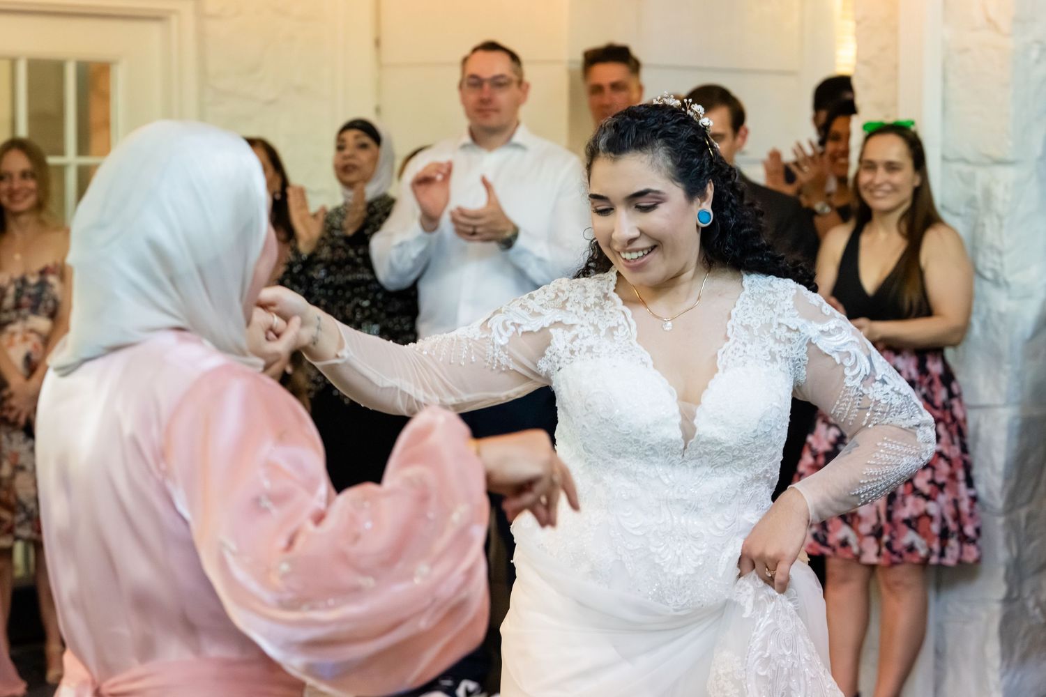 Bride in white lace gown dancing joyfully with a guest in hijab at a wedding reception.