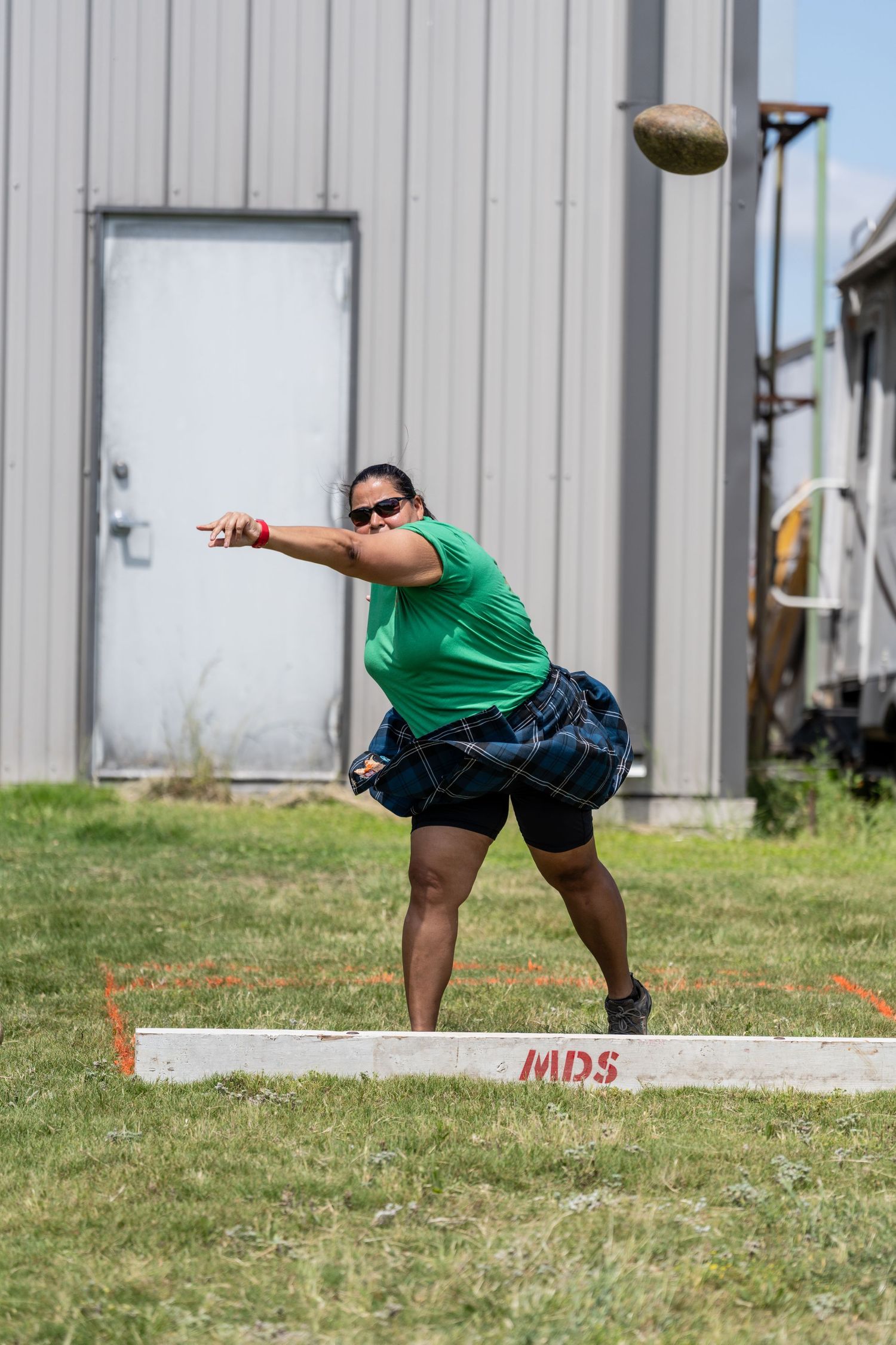 Woman in green shirt throwing a stone at a Highland Games competition outdoors near a metal building.