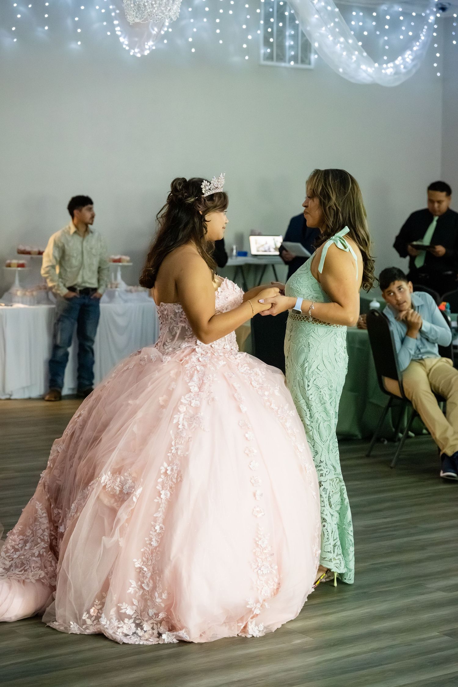Young woman in pink quinceañera ball gown and tiara shares a dance with woman in mint dress at celebration.