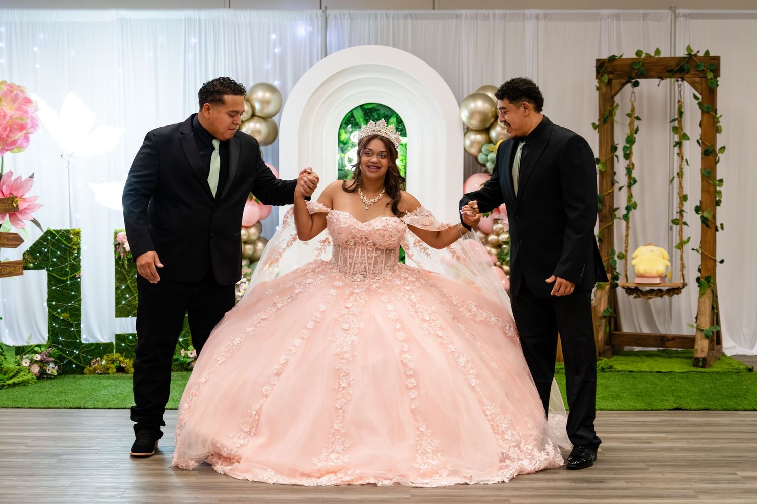 Young woman in pink quinceañera ball gown dancing with two men in suits at a decorated celebration venue.