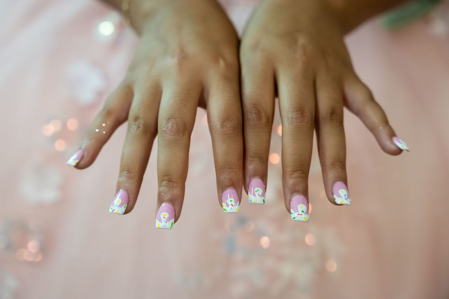 Close-up of hands with floral French tip nail art featuring pink and green flower designs with rhinestones.