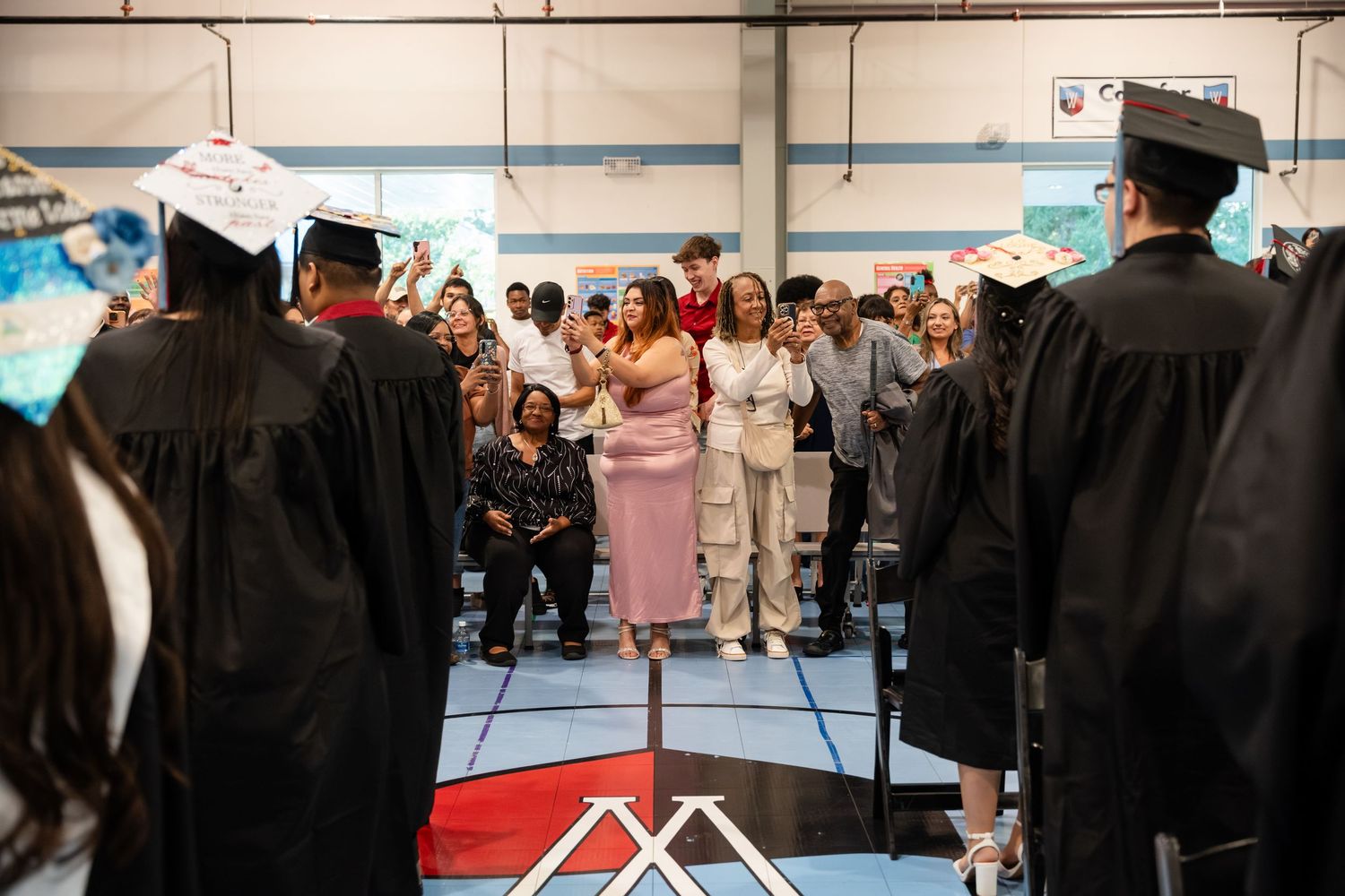 Graduation ceremony with students in caps and gowns gathered around a speaker on a gym stage.