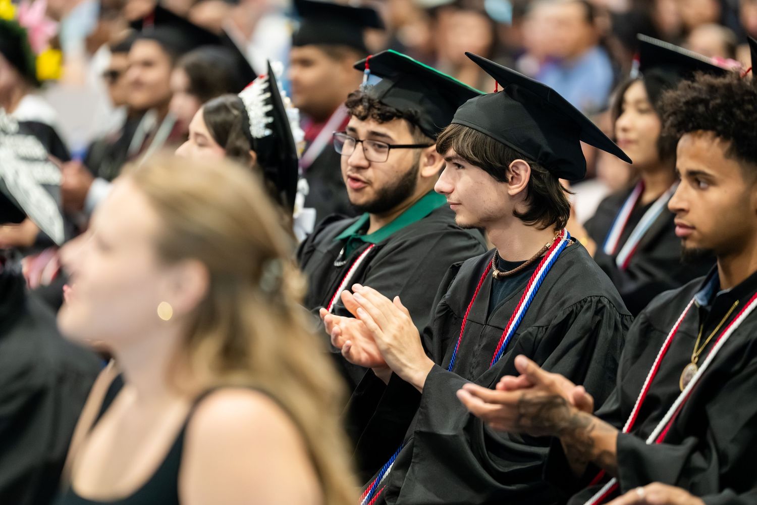 College graduates in black caps and gowns applaud during a university commencement ceremony indoors.