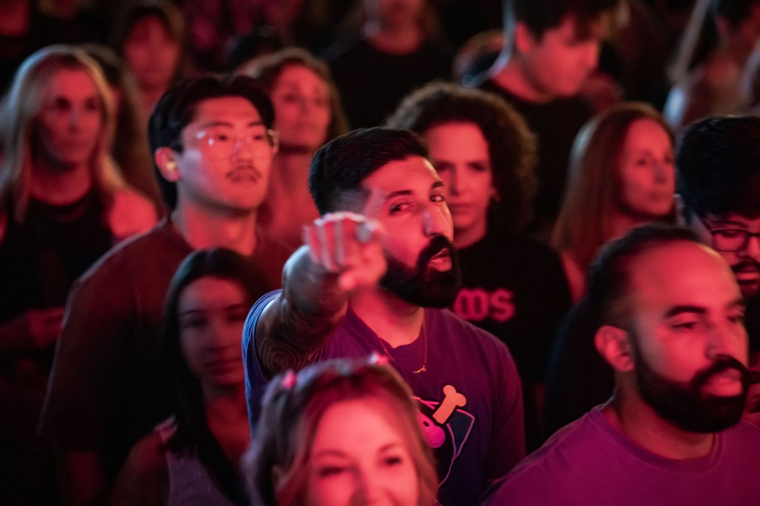A diverse crowd at a concert, with a bearded man in a purple shirt pointing forward under red stage lighting.