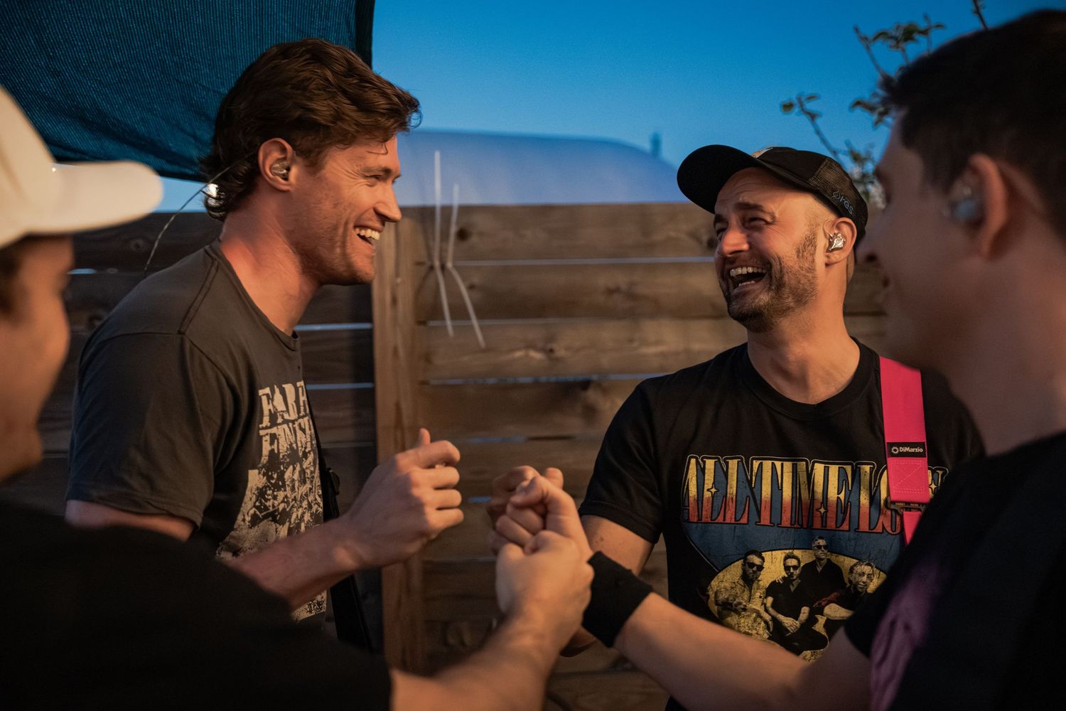 Group of friends laughing and socializing outdoors at dusk, wearing casual band t-shirts in a relaxed setting.
