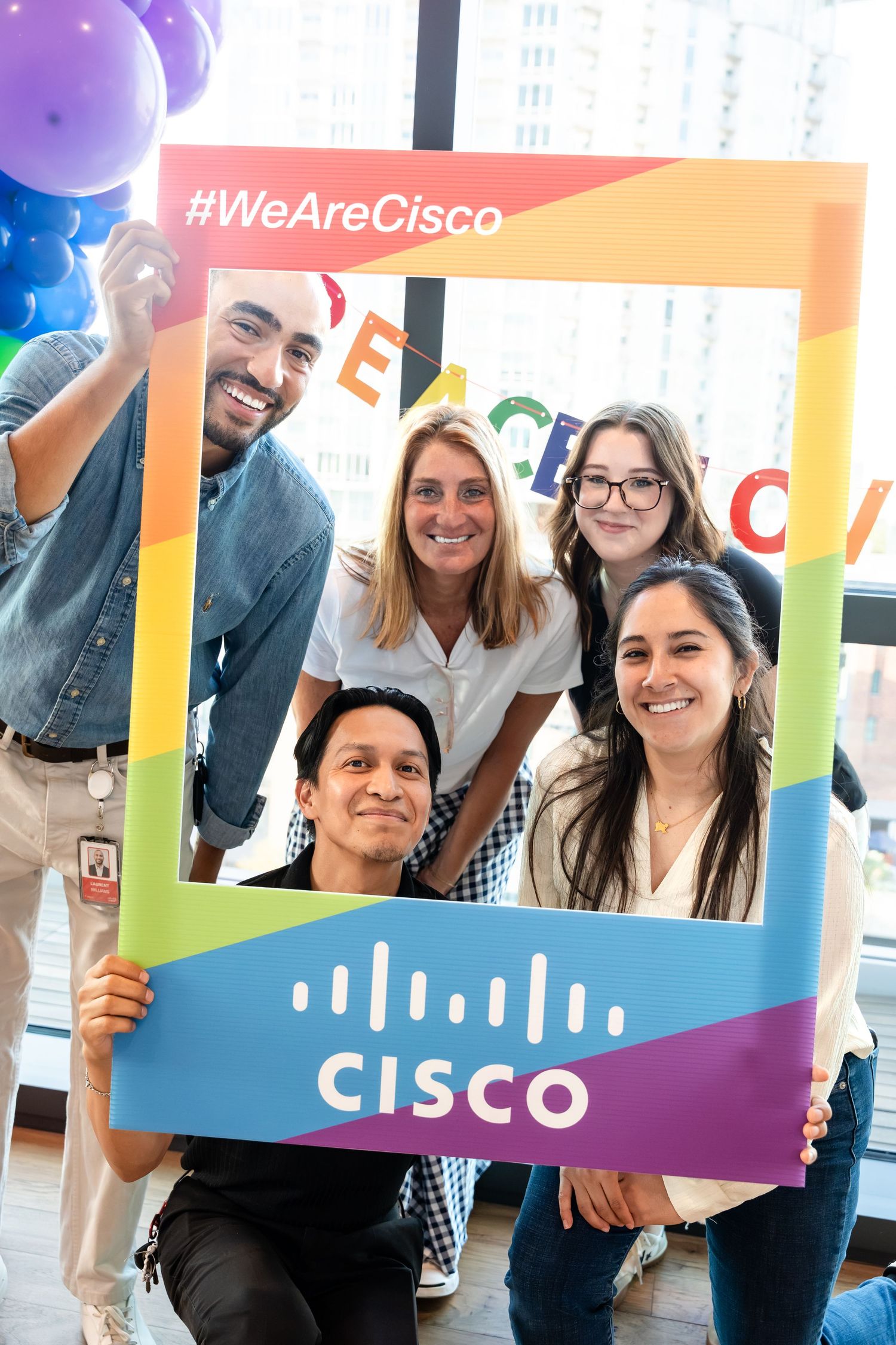 Five Cisco employees pose together holding a rainbow Pride photo frame with #WeAreCisco hashtag at a corporate event.