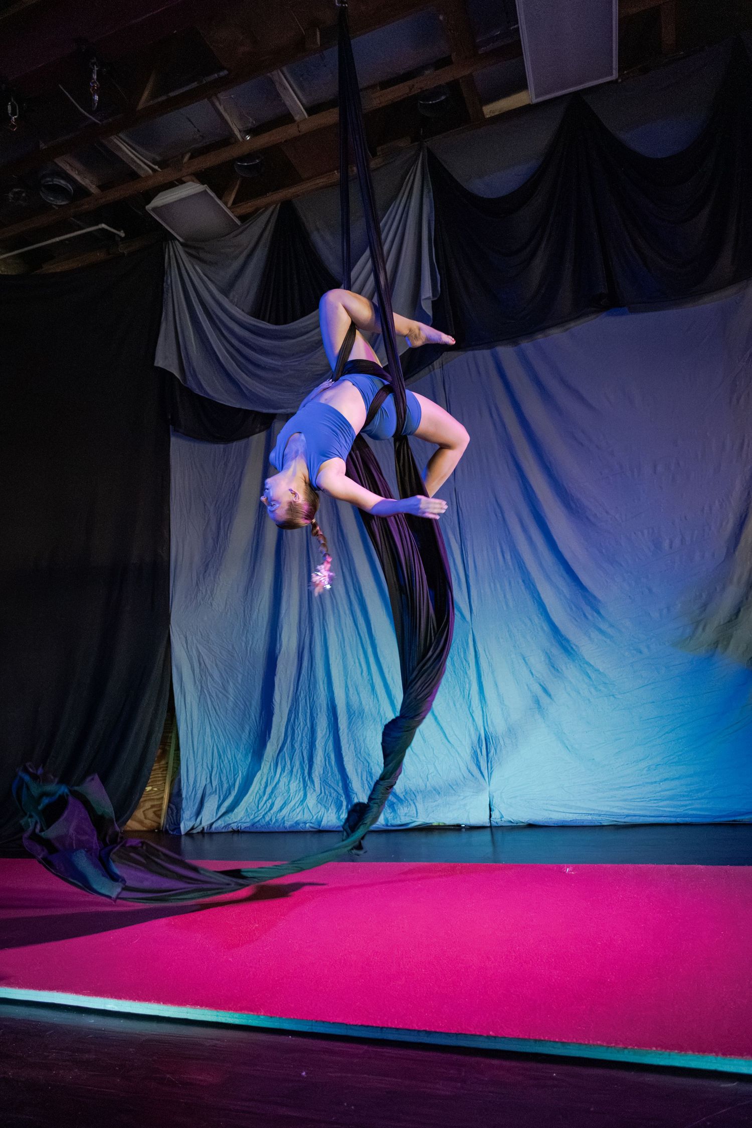 Aerial silk performer in blue outfit executing an inverted pose on stage with pink floor and dark draped backdrop.