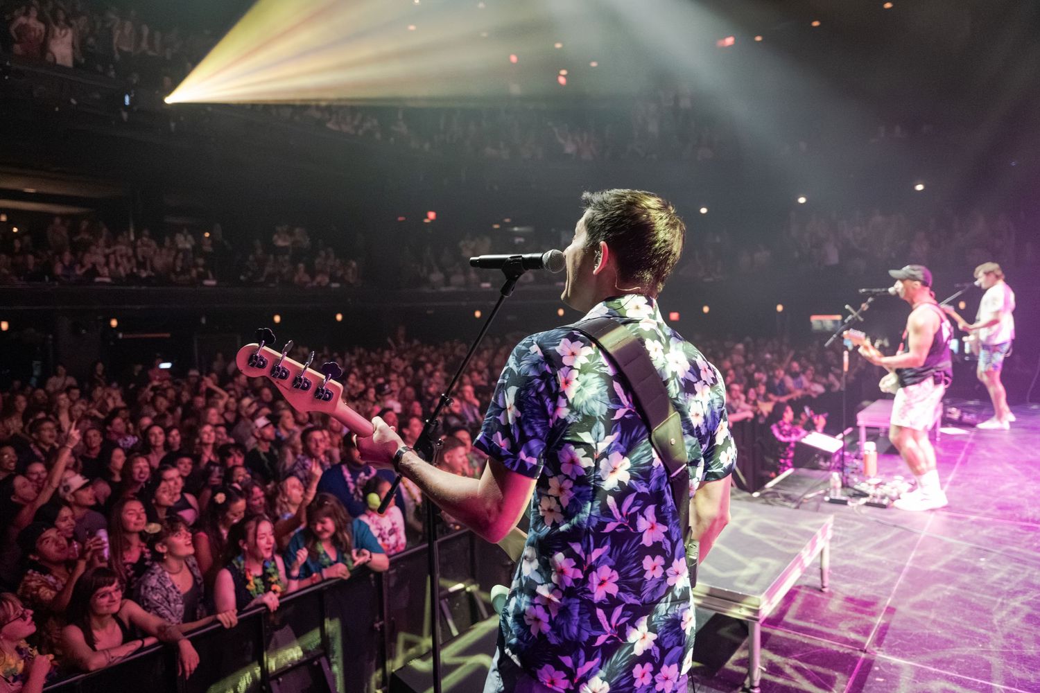Performer in floral shirt sings to packed concert crowd under colorful stage lights at live music venue.