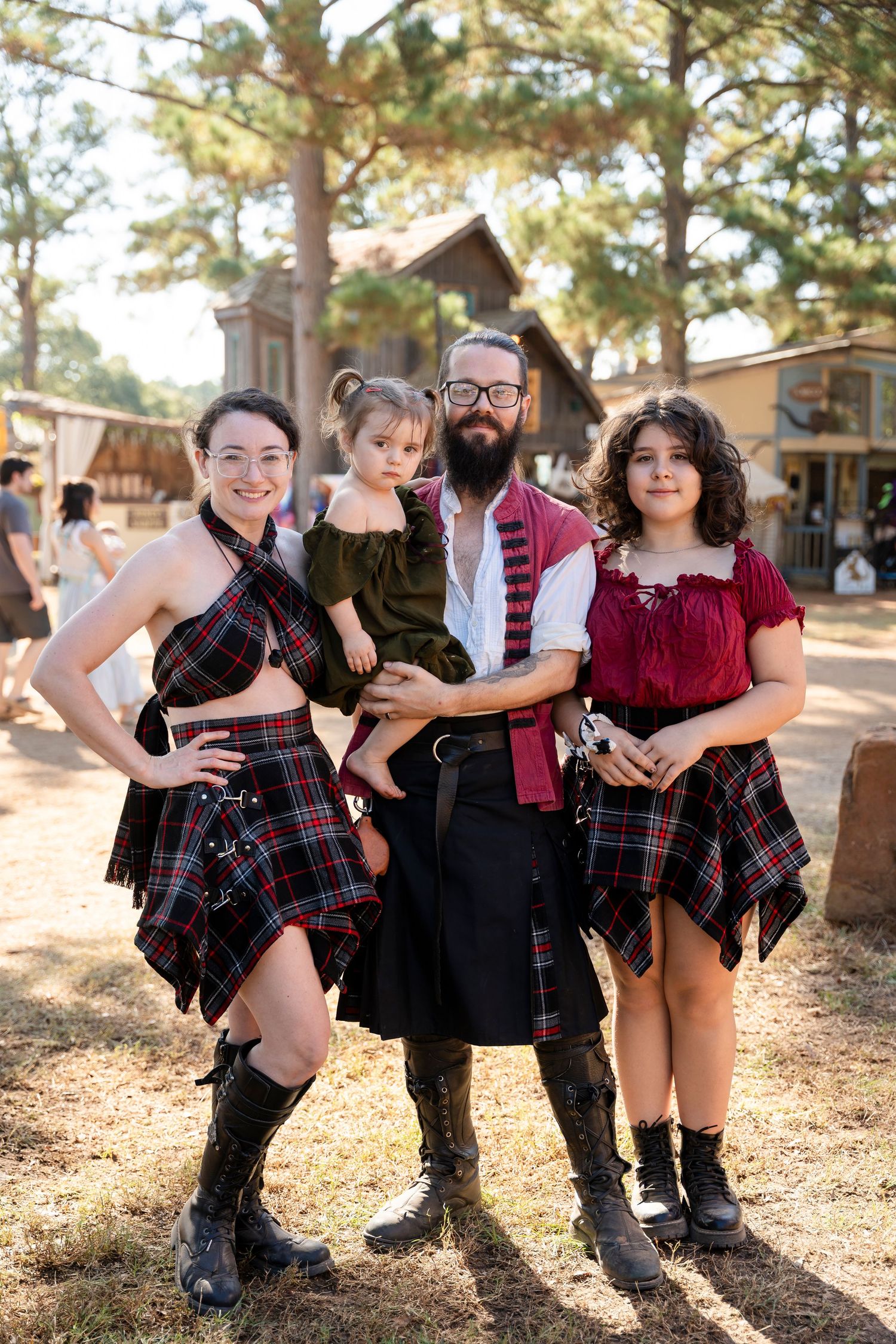 A group wearing coordinated plaid and burgundy outfits poses playfully in a sunlit outdoor setting among pine trees.