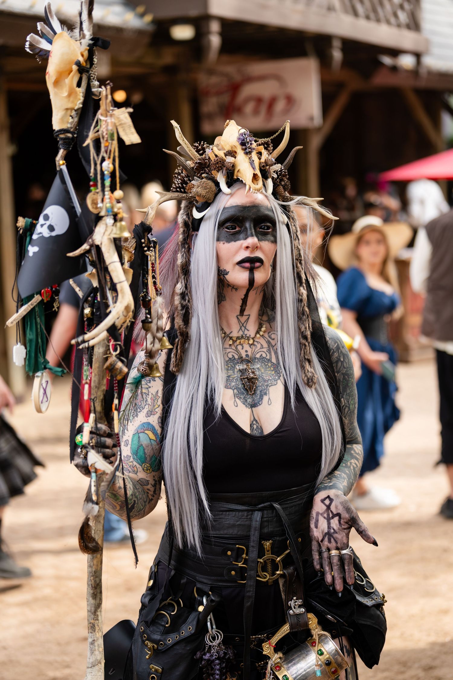A costumed figure in black attire with long gray hair and face paint stands at a medieval renaissance festival.