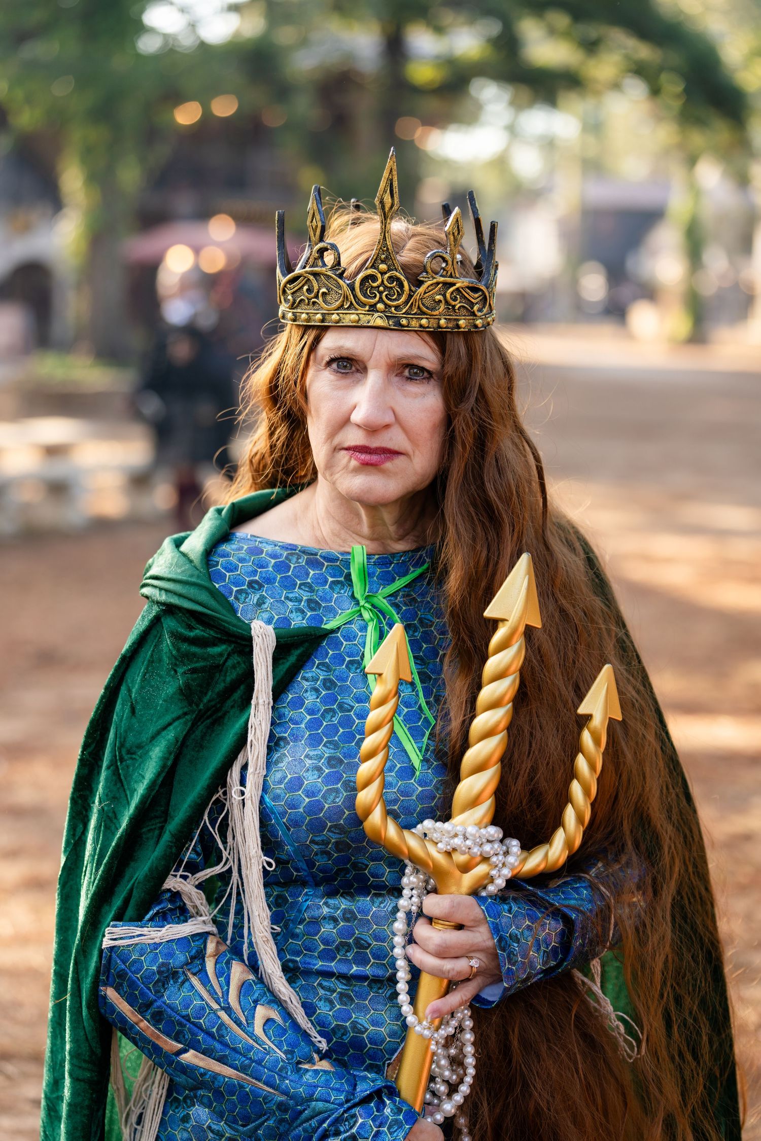Medieval cosplayer in emerald green cloak and blue dress holds golden twisted candles while wearing ornate crown.