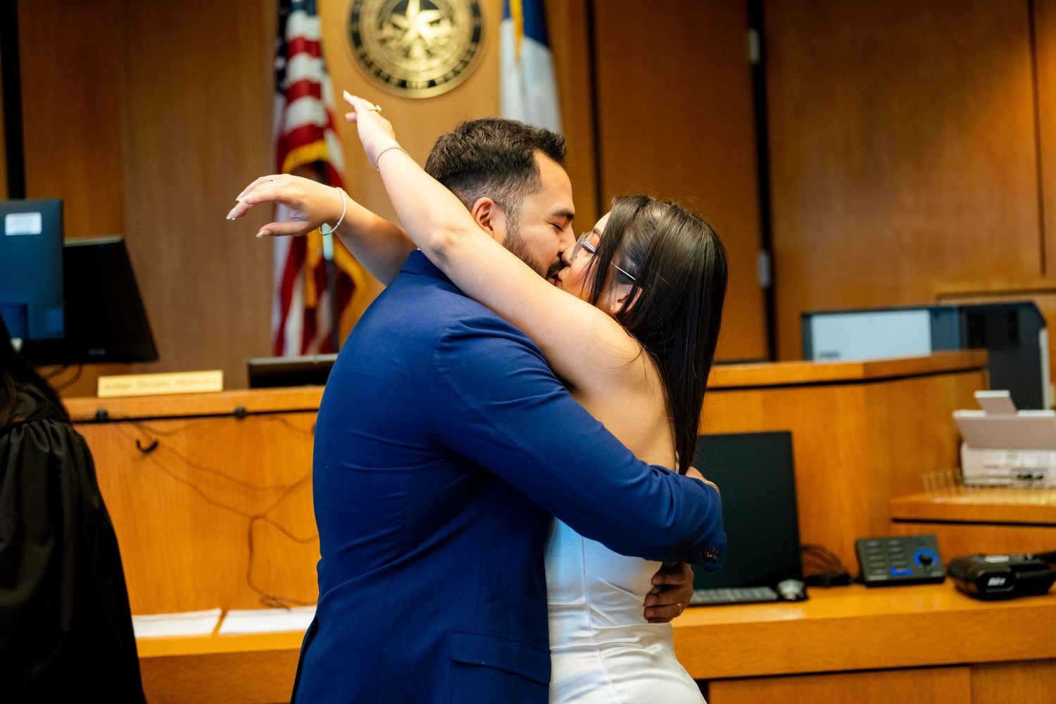 Two people embrace emotionally in a courtroom with an American flag visible in the background.