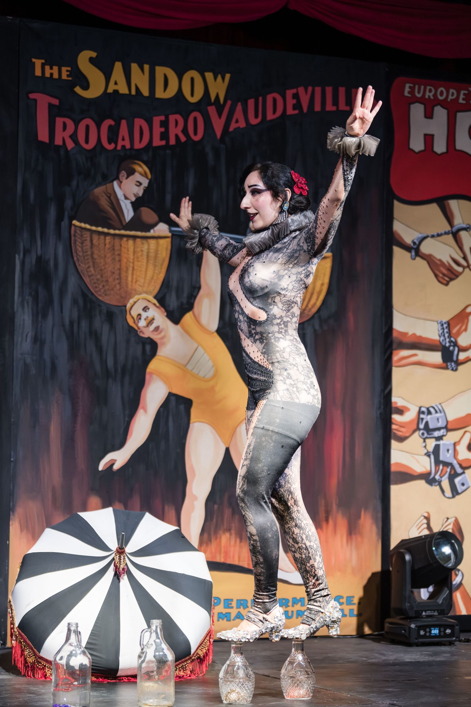 A performer in a silver costume strikes a dramatic pose on stage with vintage vaudeville-style backdrop and props.