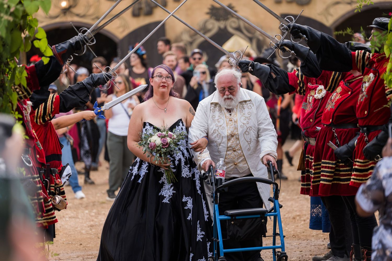 A couple walks through a medieval-themed celebration with costumed performers in red outfits lining their path.