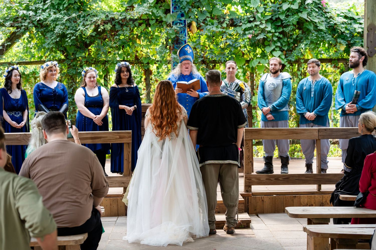 Outdoor wedding ceremony with wooden benches arranged in rows under a shaded canopy of trees.