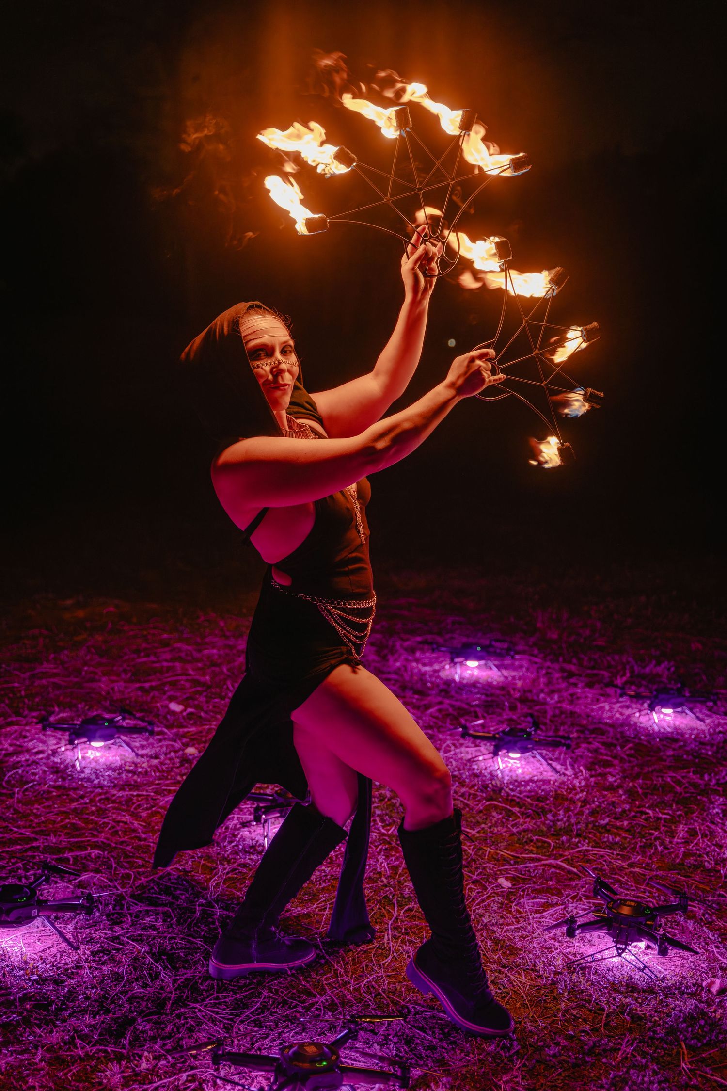 A female fire dancer performs at night, spinning flaming props under purple lights in a dramatic outdoor show.