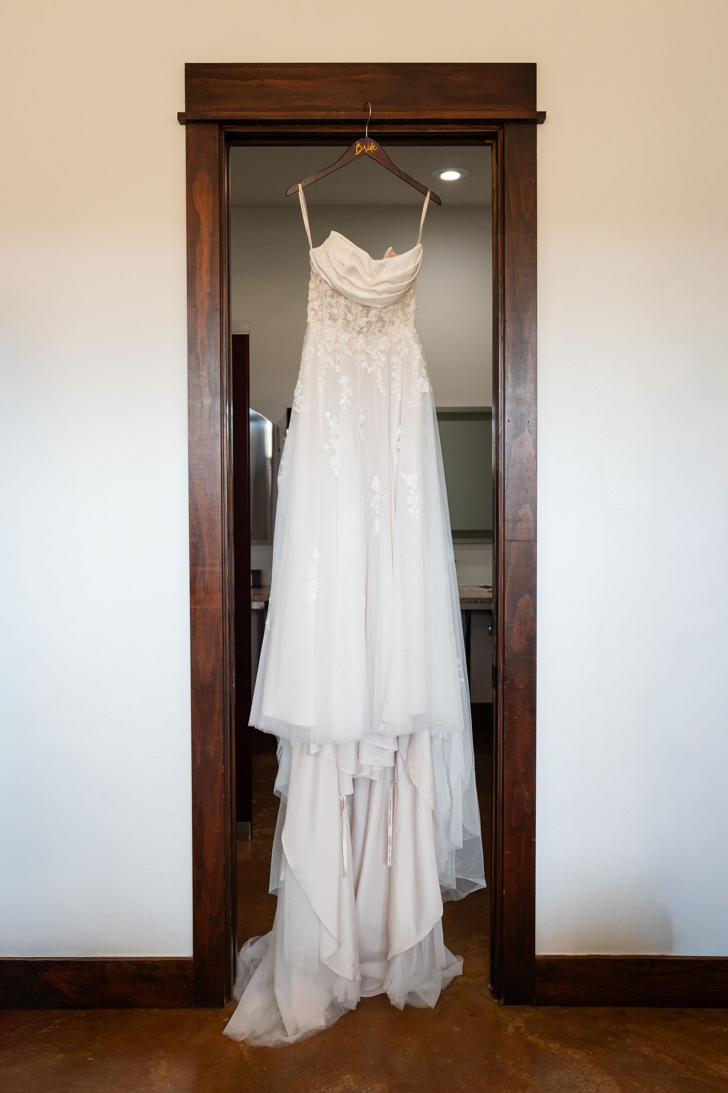 Elegant white wedding dress with lace bodice hanging on a wooden door frame in a bright room.