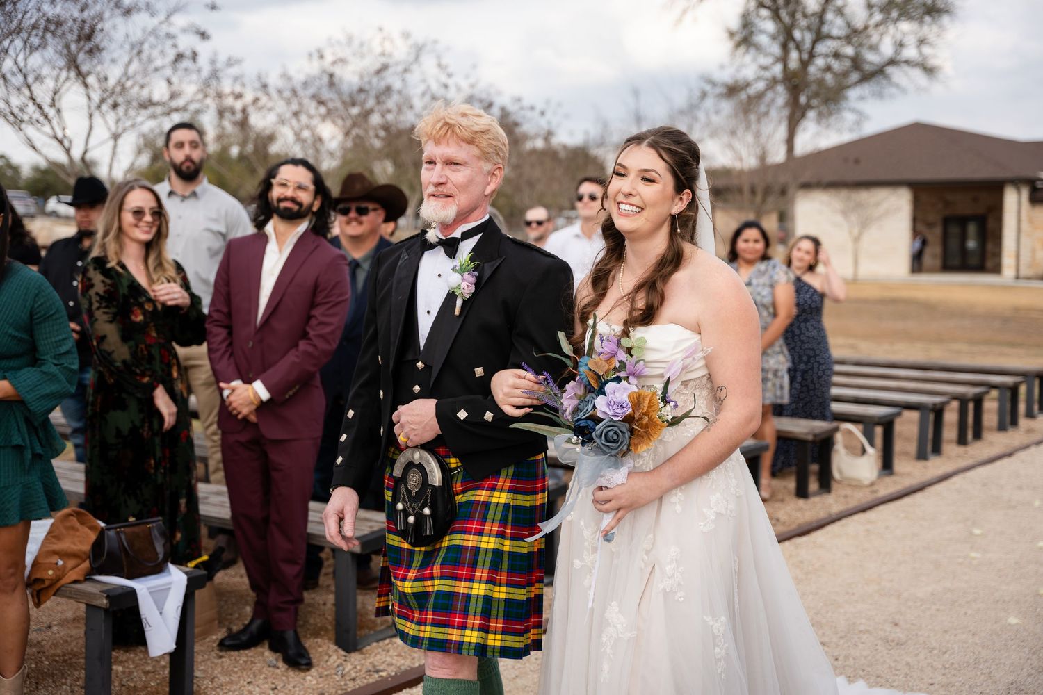 Bride in white gown and groom in colorful Scottish kilt walk down outdoor wedding aisle with guests watching.