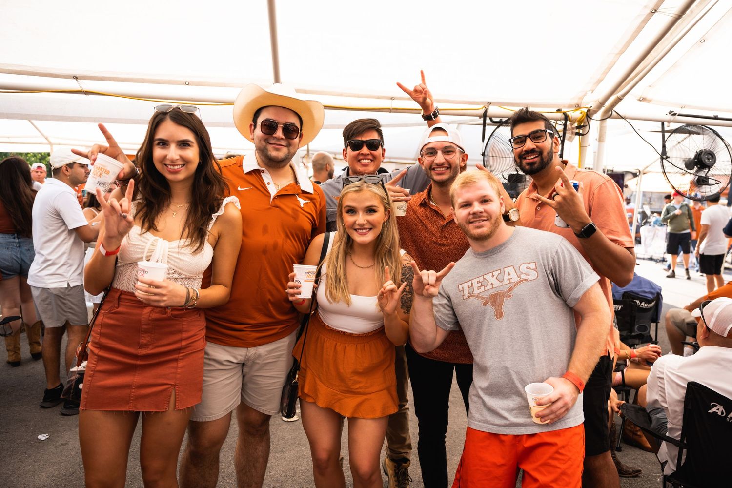 A group of friends in burnt orange and white outfits celebrating together under a white tent at an outdoor event.