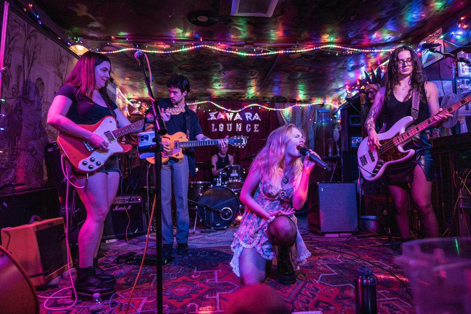 Musicians perform on stage at a dimly lit venue with colorful purple and blue lighting and string lights overhead.