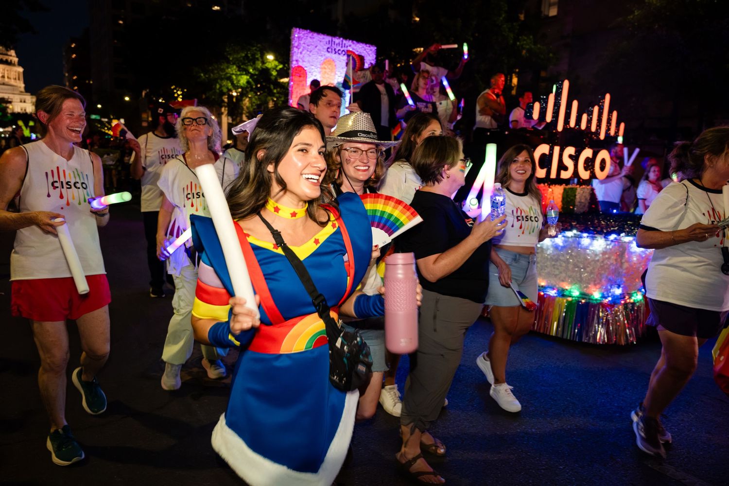 Cisco employees celebrate Pride parade at night, wearing colorful rainbow accessories and dancing near illuminated Cisco float.