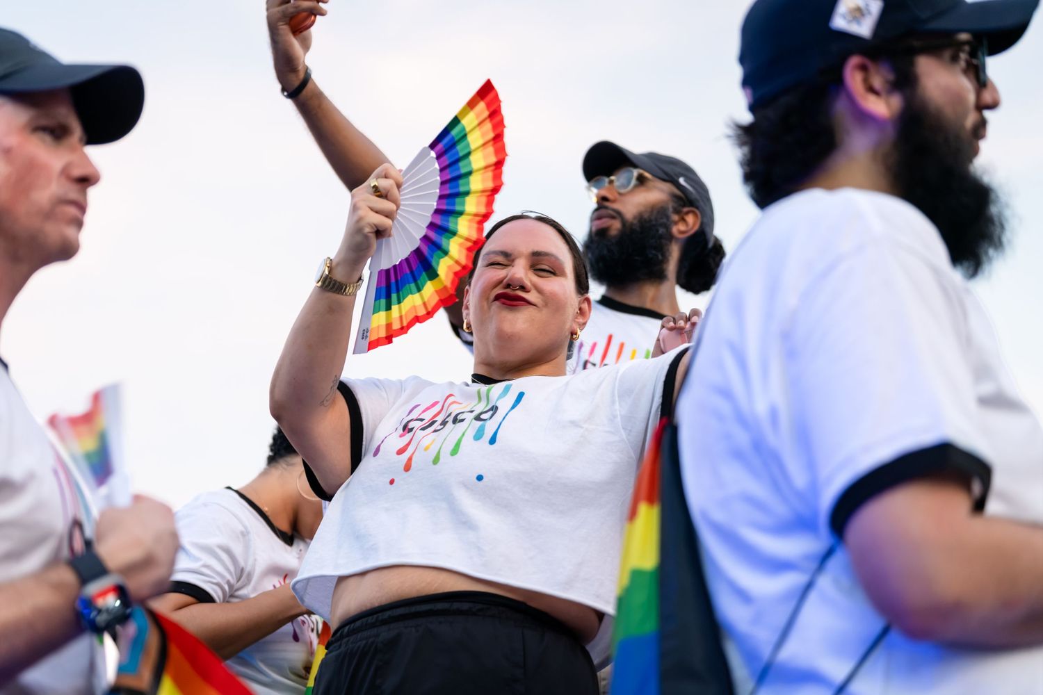 Woman holding rainbow fan at Pride parade with diverse crowd wearing rainbow accessories in background.