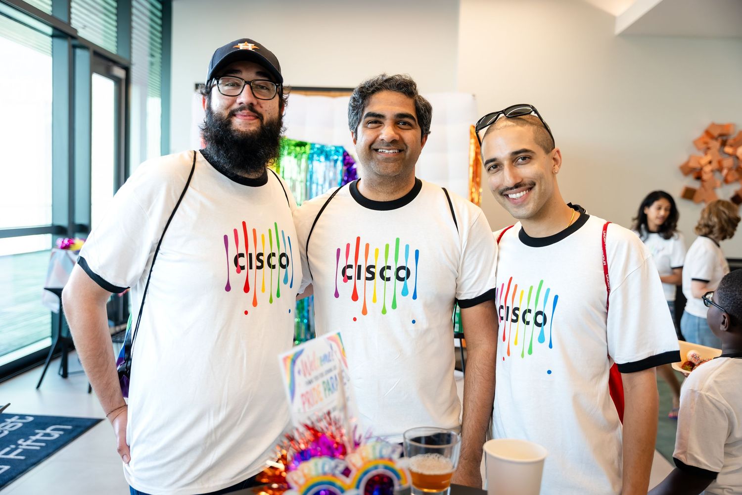 Three men wearing Cisco pride t-shirts smile together at a colorful corporate event.