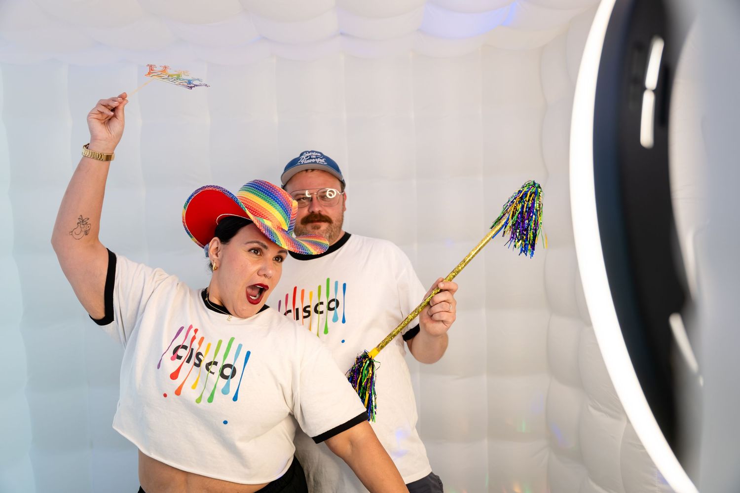 Two people in Cisco Pride shirts pose energetically in a photo booth, wearing rainbow accessories and holding colorful pompoms.