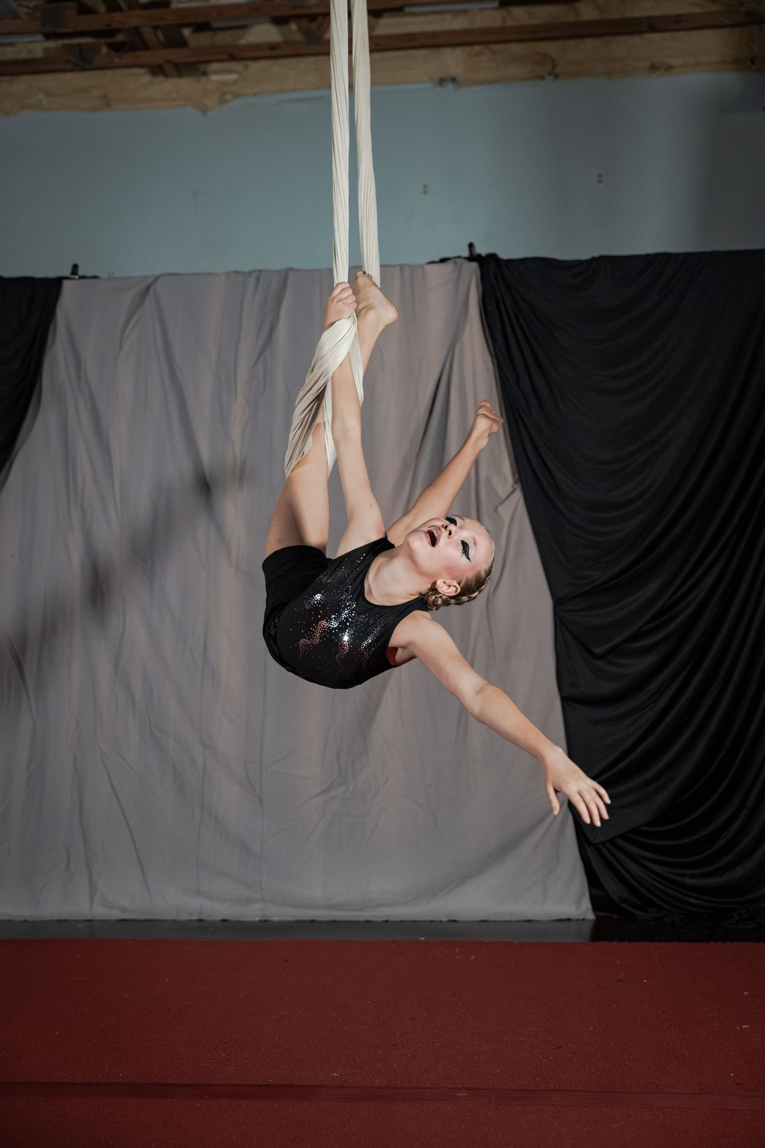 Aerial silk performer in black sequin leotard gracefully poses mid-air on white silks in a studio setting.