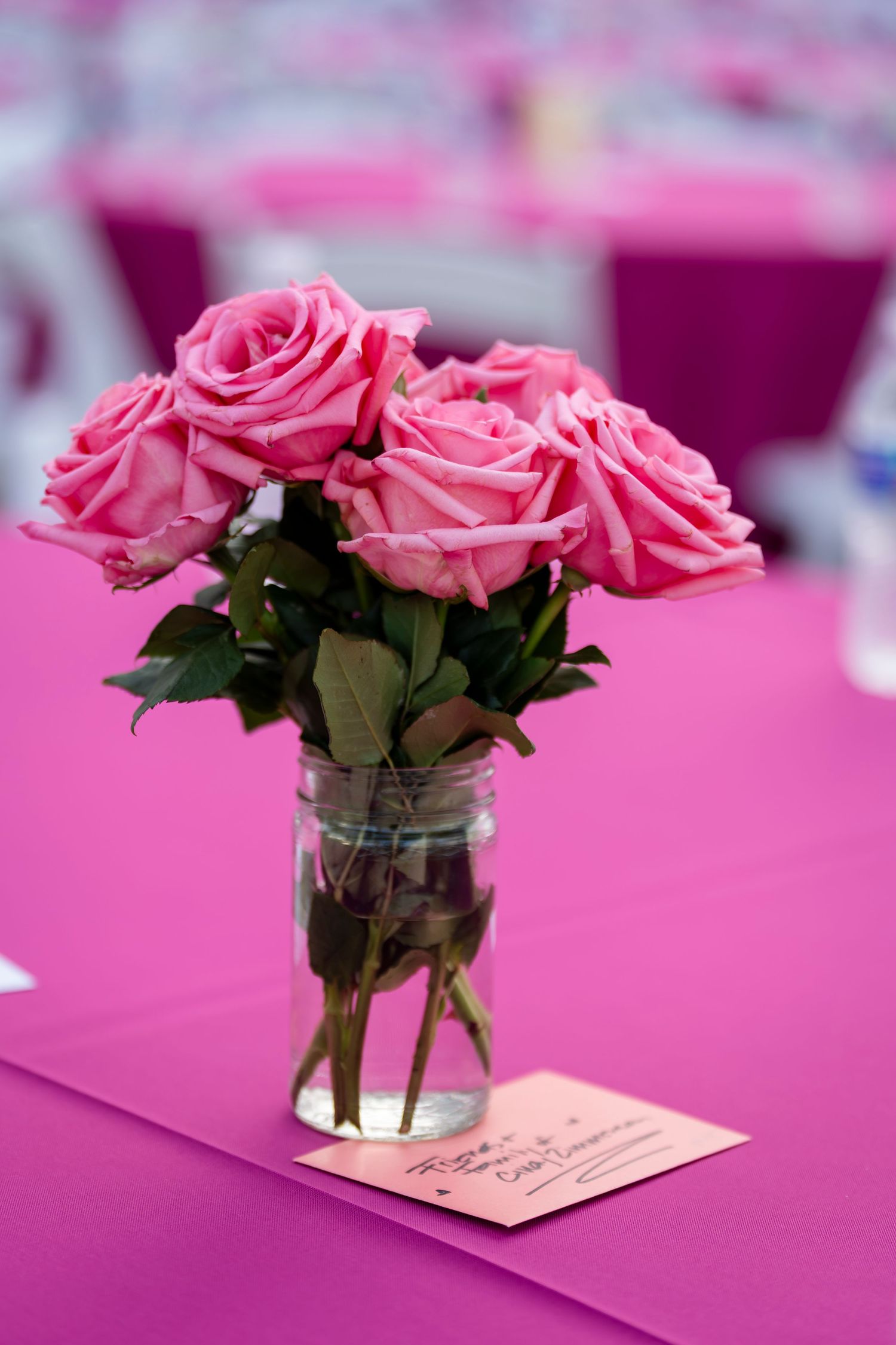 Pink roses in a glass mason jar vase on a bright pink tablecloth with a small card beside it.