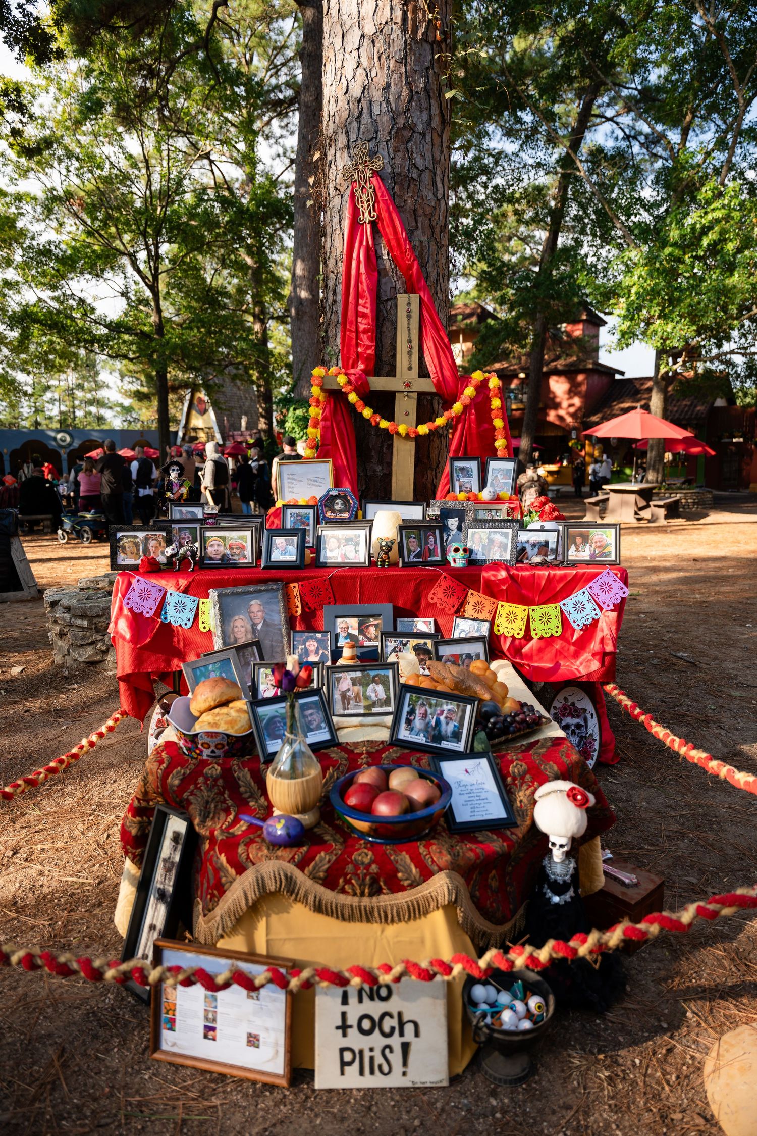 A colorful outdoor memorial display with photos, red decorations, and personal items arranged on wooden platforms in a park setting.