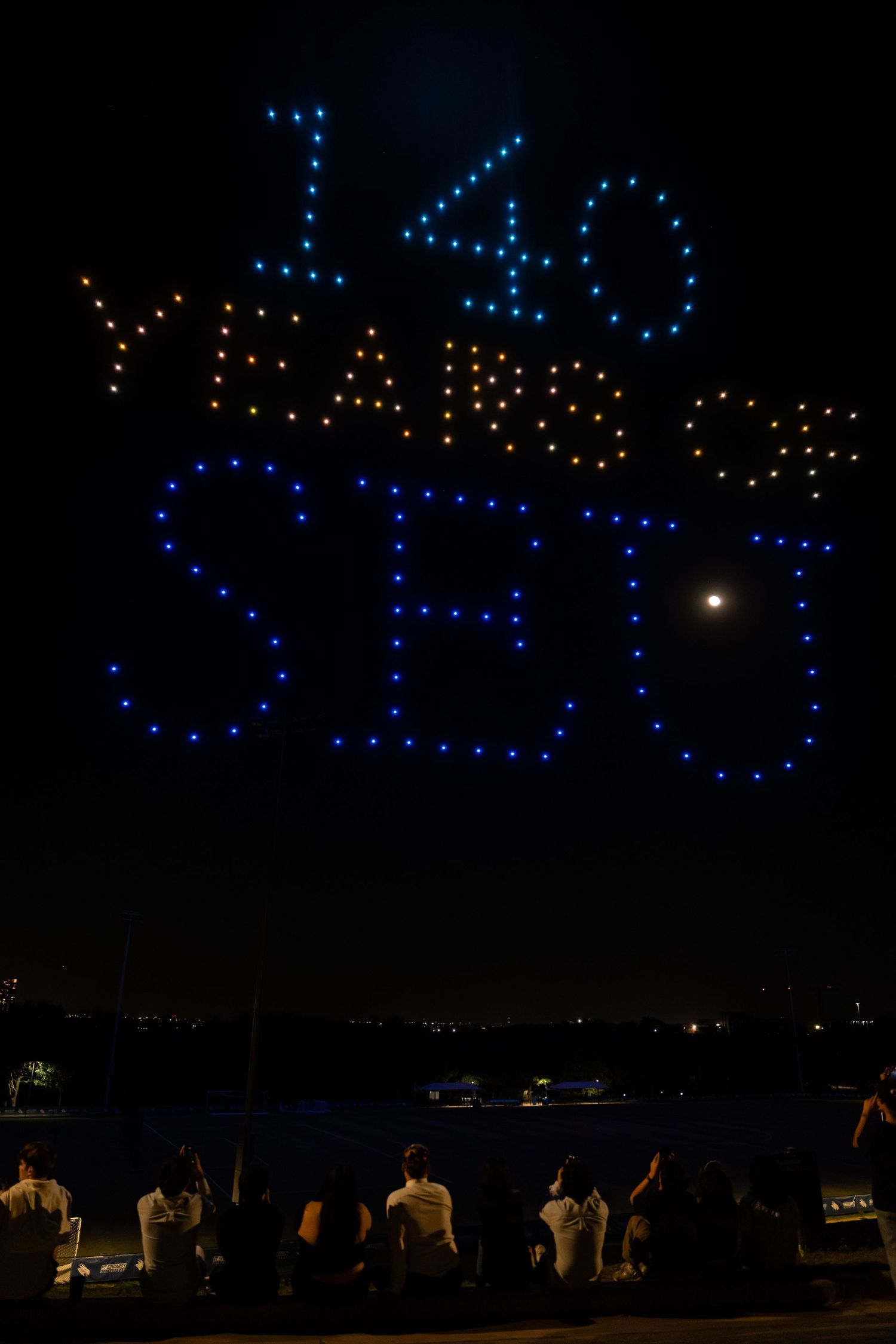 Illuminated drone light show displaying '140' in blue and orange lights against night sky as spectators watch from below.