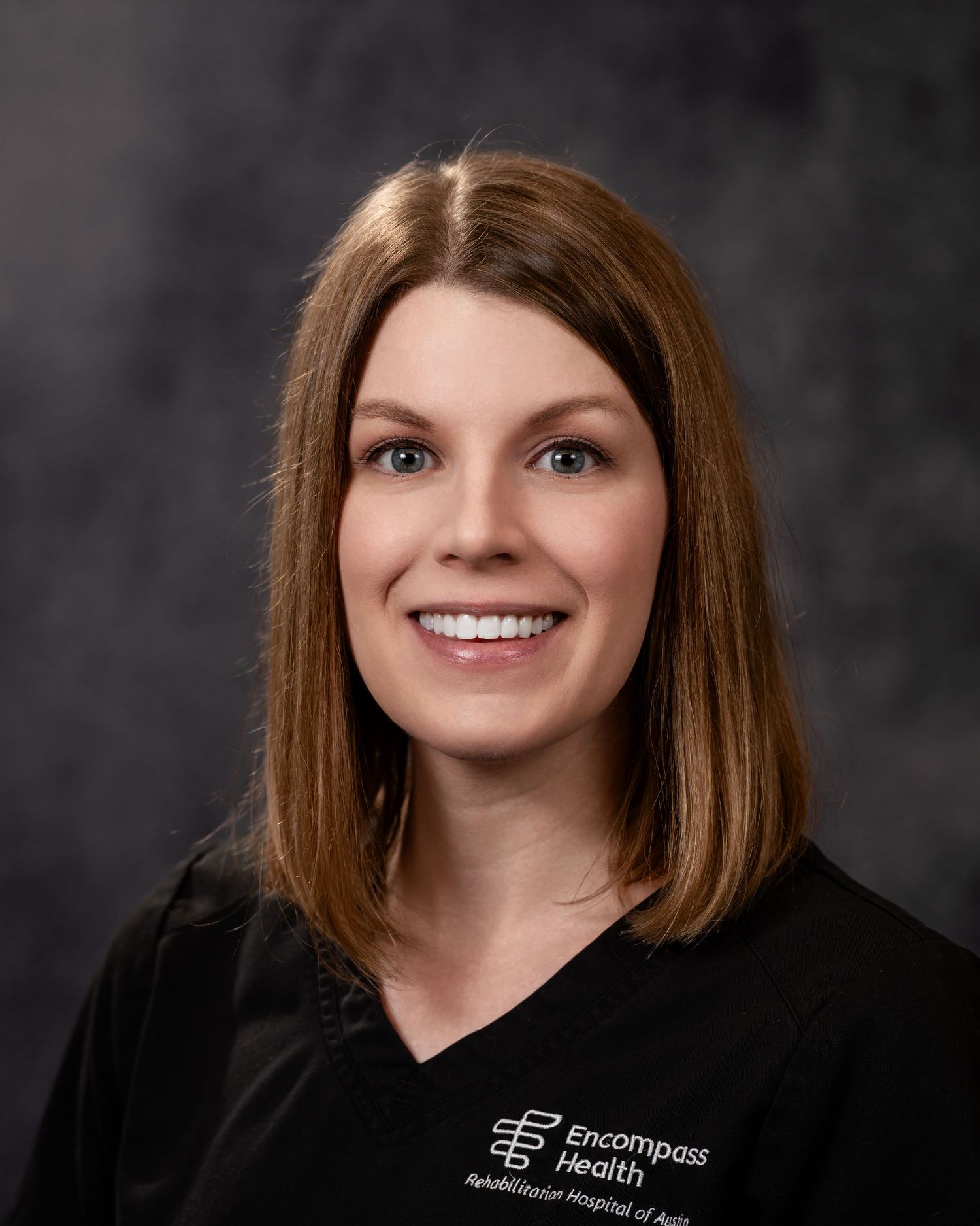 Professional headshot portrait of a healthcare worker wearing a black uniform with Encompass Health logo against gray background.