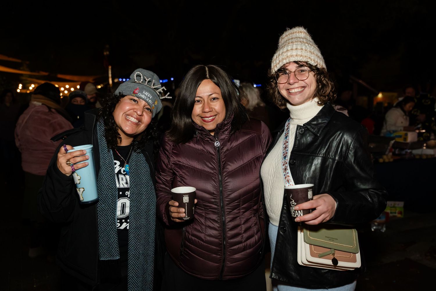 Friends gather together at a nighttime outdoor event holding drinks and smiling for the camera.