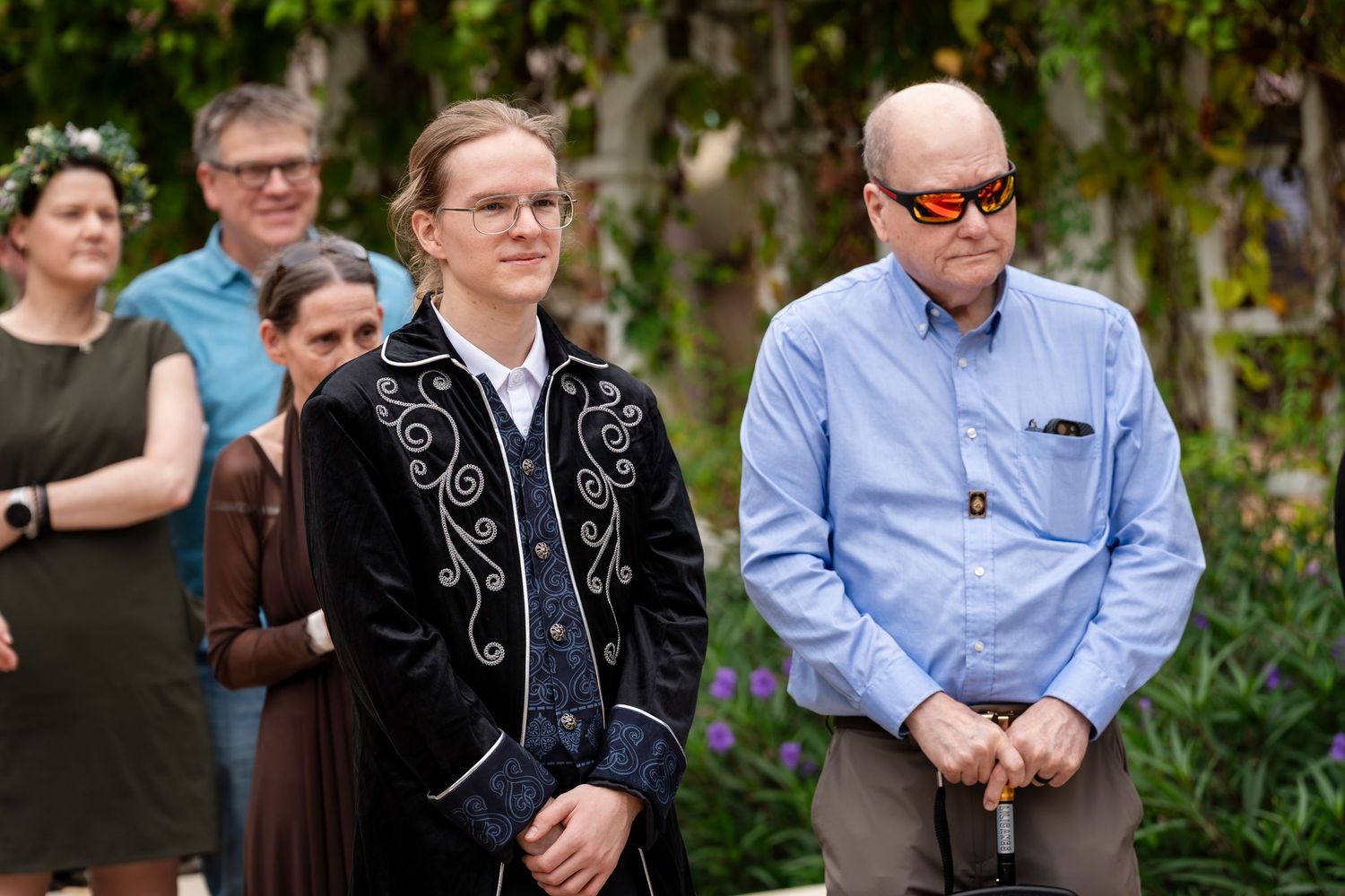 Two people stand outdoors, one wearing an embroidered black jacket and another in a light blue button-down shirt.
