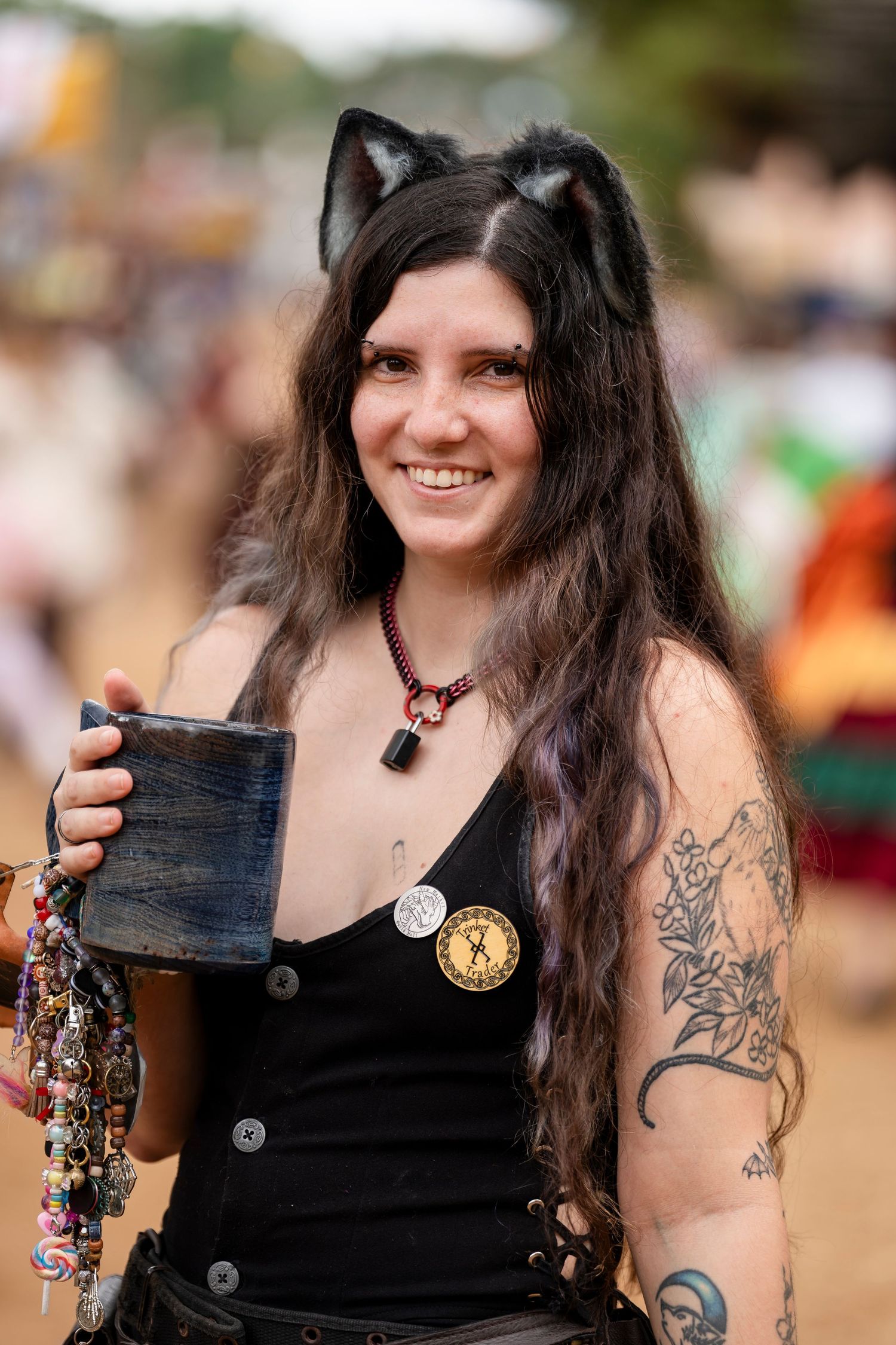 Person with long dark hair and tattoos wearing a black tank top holds up a decorative drinking vessel at a festival.