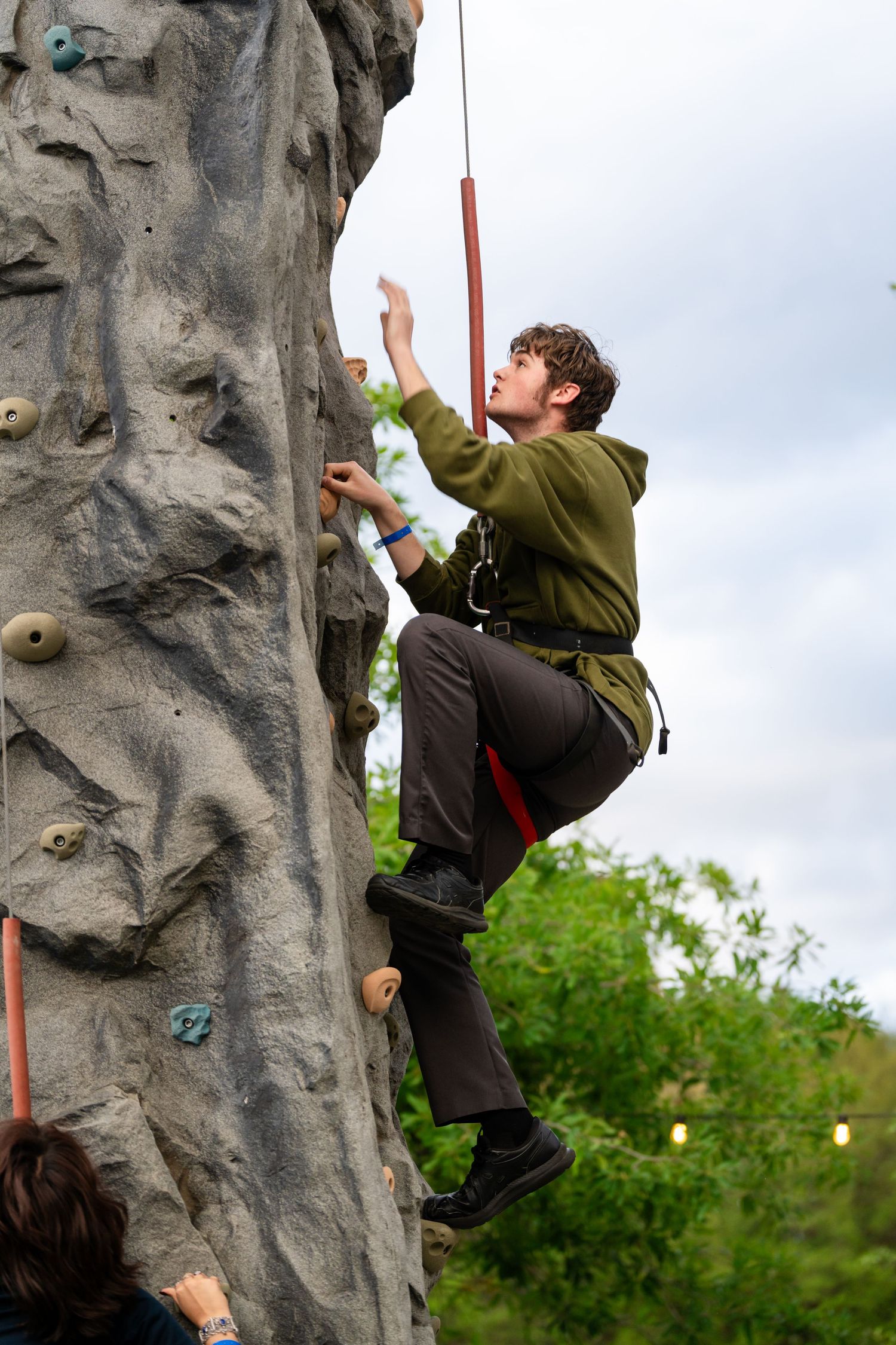 Young man rock climbing on an outdoor climbing wall, wearing a green hoodie and safety harness.