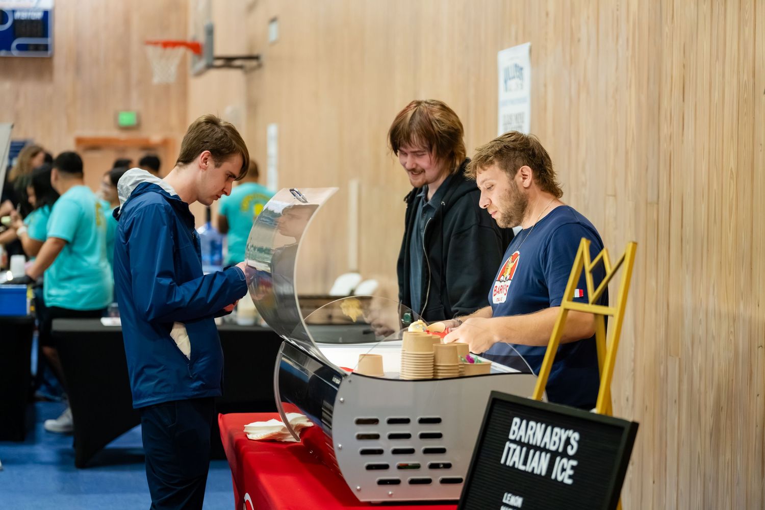 Three young men at a Barnaby's Italian Ice stand during an indoor food event in a school or community center.